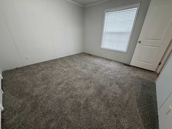 Empty room with light gray walls, brown carpet, a closed white door, and a large window with white blinds. The room is clean, simple, and well-lit with natural light.