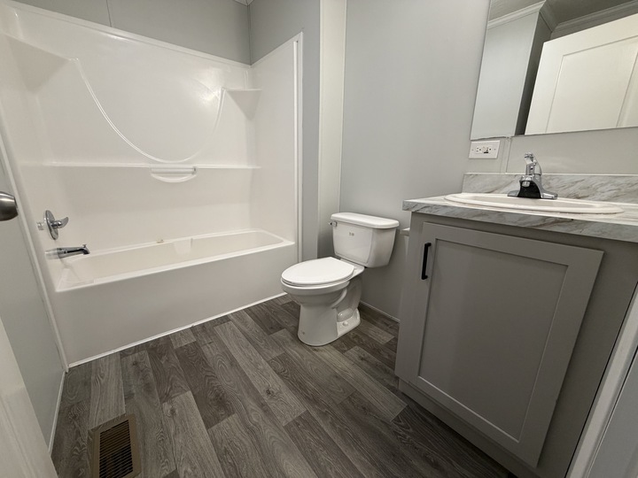 A modern bathroom with gray wood-look flooring, a white bathtub-shower combo, a white toilet, and a gray vanity with a marble countertop and sink beneath a mirror. The walls are painted light gray.