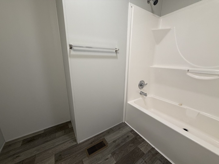 A clean, modern bathroom with white walls, a towel bar, a white bathtub with built-in shelves, chrome fixtures, and wood-look vinyl flooring. An air vent is visible on the floor.