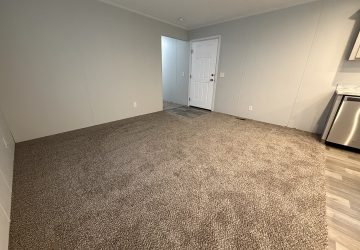 A room with beige carpet, gray walls, and white trim. Theres a white door on the far wall, a small section of tile flooring near the entrance, and part of a stainless steel appliance is visible on the right.