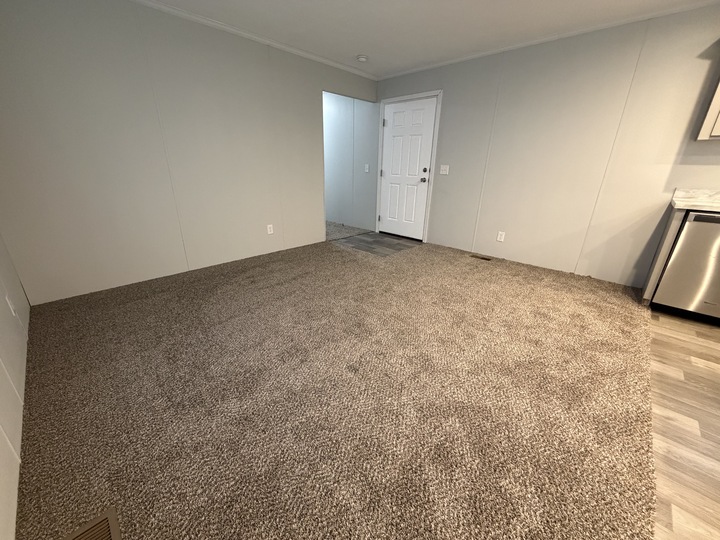 A room with beige carpet, gray walls, and white trim. Theres a white door on the far wall, a small section of tile flooring near the entrance, and part of a stainless steel appliance is visible on the right.