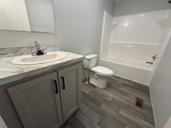 A modern bathroom with gray cabinets, a marble countertop sink, a white toilet, and a white bathtub-shower combo. The floor has gray wood-style planks, and the walls are light gray.