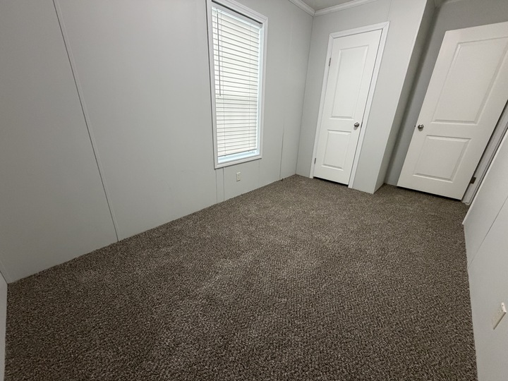 Empty room with light gray walls, a window with closed white blinds, two white doors, and brown carpet flooring. The room is well-lit and appears clean and unfurnished.