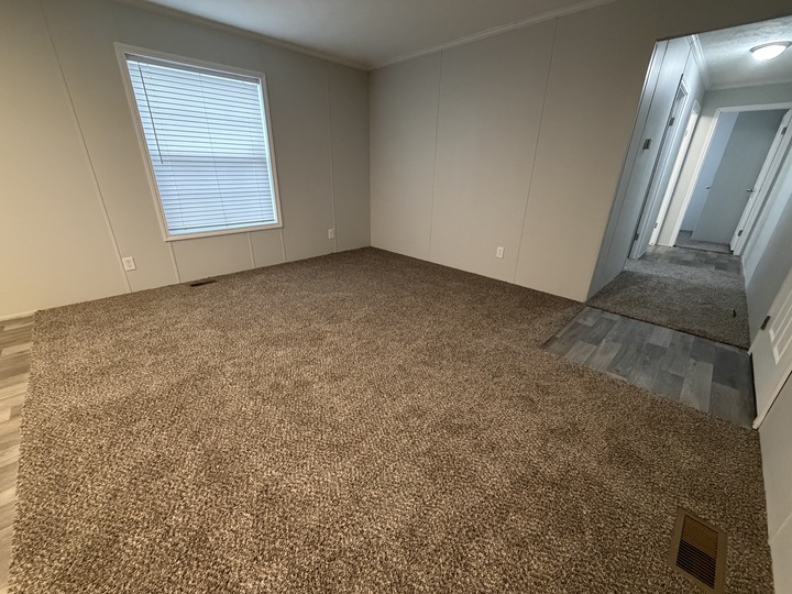 Empty room with beige carpet, light gray walls, a single window with white blinds, and a hallway with gray wood-look flooring. The room appears clean and unfurnished.