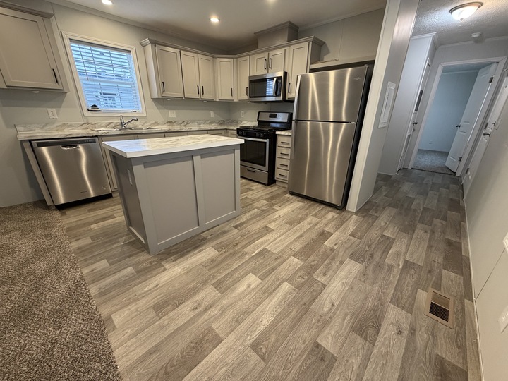 Modern kitchen with gray cabinets, stainless steel appliances, a central island with a marble countertop, wood-look flooring, and carpeted area leading to a hallway with several doors.