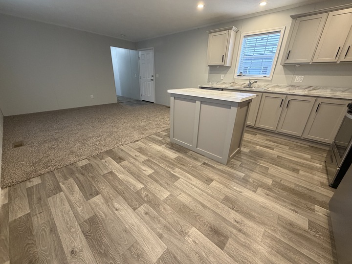 Modern open-concept room with a kitchen featuring light gray cabinets, marble countertops, and wood-look flooring. Adjacent living area is carpeted, with a window, entry door, and neutral wall colors.