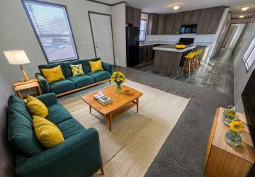 A modern open-plan living room and kitchen with teal sofas, yellow cushions, a wooden coffee table with flowers, a sideboard with vases, and a kitchen island with yellow barstools and a pumpkin on top.