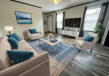 A modern living room with a beige sofa, two matching armchairs with teal pillows, a patterned blue rug, a coffee table, a flat-screen TV on the wall, large windows with curtains, and a potted plant in the corner.
