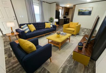 A modern living room with a navy blue sofa, matching armchair, a yellow armchair, and a yellow ottoman around a coffee table. A TV sits on a wooden stand. The space opens to a kitchen in the background.