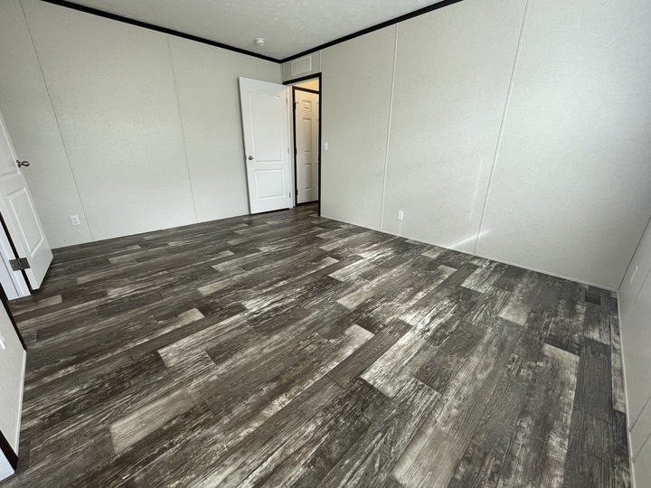 Empty room with light-colored walls, dark trim, and wood-patterned vinyl flooring. The door is open, revealing a small hallway and another closed door beyond. The room is well-lit by natural light.