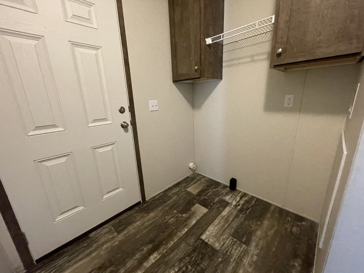 A small, empty laundry room with a wooden floor, a white door, two brown wall cabinets, a wire shelf, and a wall outlet with washer/dryer hookups. The room has beige walls and is well-lit.