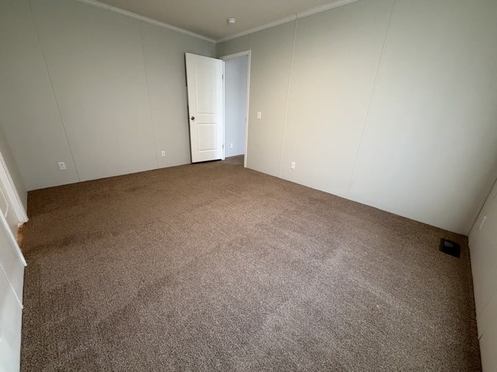 Empty room with beige carpet, light gray walls, white trim, and a white door partially open. The room has no furniture, a ceiling vent, and appears clean and well-lit.