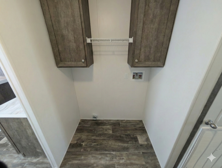 A small laundry area with wood-look flooring, two upper cabinets, a wire shelf, and hookups for a washer and dryer on a white wall. A doorway leads into an adjacent room.