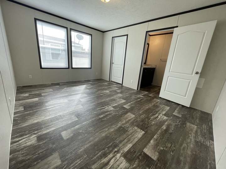 A spacious, empty room with dark wood-look flooring, two large windows, and two open white doors—one leading to a closet and the other to a bathroom with a visible sink.