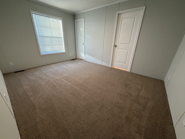 A small, empty room with beige carpet, light gray walls, a window with closed blinds, and two white doors. Sunlight streams through the window, casting shadows on the floor.