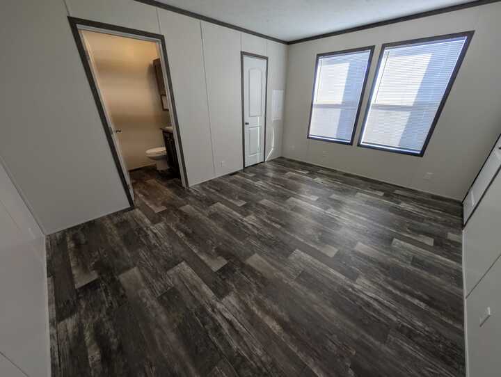 A bright, empty room with dark wood flooring, white walls, large windows with blinds, a white door, and an open doorway leading to a bathroom with a visible toilet.