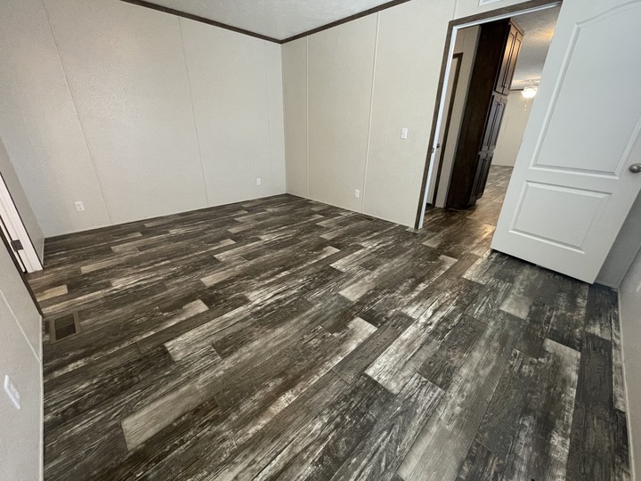 Empty room with light beige walls, dark brown trim, and grayish wood-look vinyl flooring. An open door leads to a hallway with similar flooring and visible cabinetry in the adjoining space.
