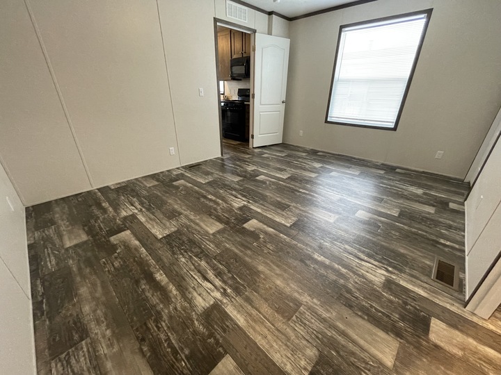 A mostly empty room with wood-style vinyl flooring, beige walls, a large window with blinds, an open white door, and a view into a kitchen with dark cabinets.