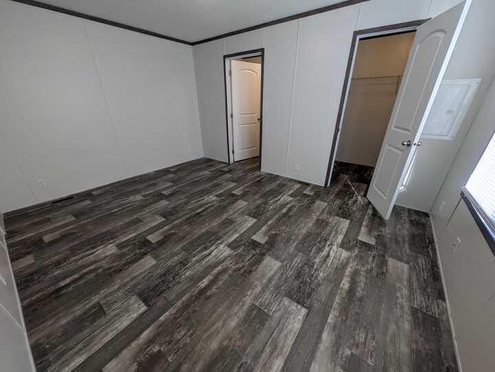 Empty room with gray wood-patterned flooring, white walls, and dark trim. Two open doors reveal a closet and another room. A window on the right lets in natural light.