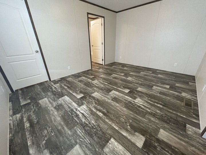 Empty room with light-colored walls, dark trim, and wood-patterned vinyl flooring in varying shades of gray and brown. Two white doors are visible, one open and one closed. The room is unfurnished.