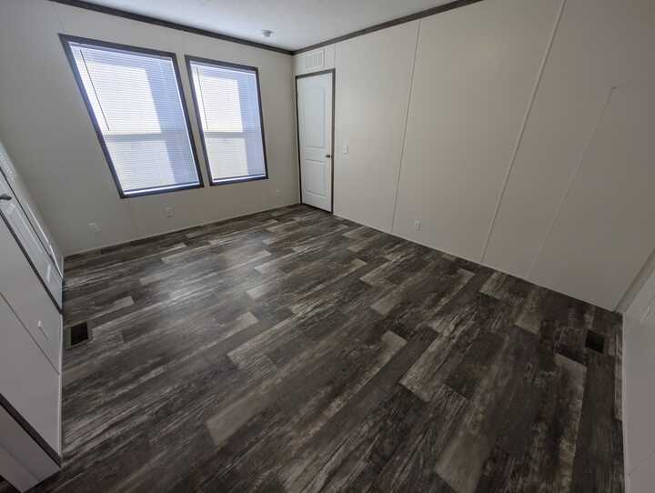 Empty room with two large windows, wood-look vinyl flooring, white walls with dark trim, and a closed white door. Natural light fills the space.