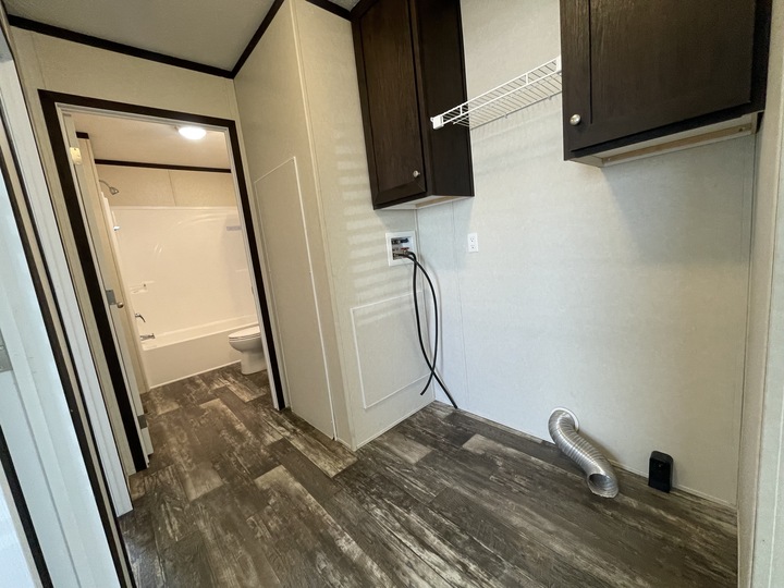A laundry room with brown cabinets, a white wire shelf, washer and dryer hook-ups, and a vent hose on a wood-look floor. A bathroom with a tub and toilet is visible through an open doorway.