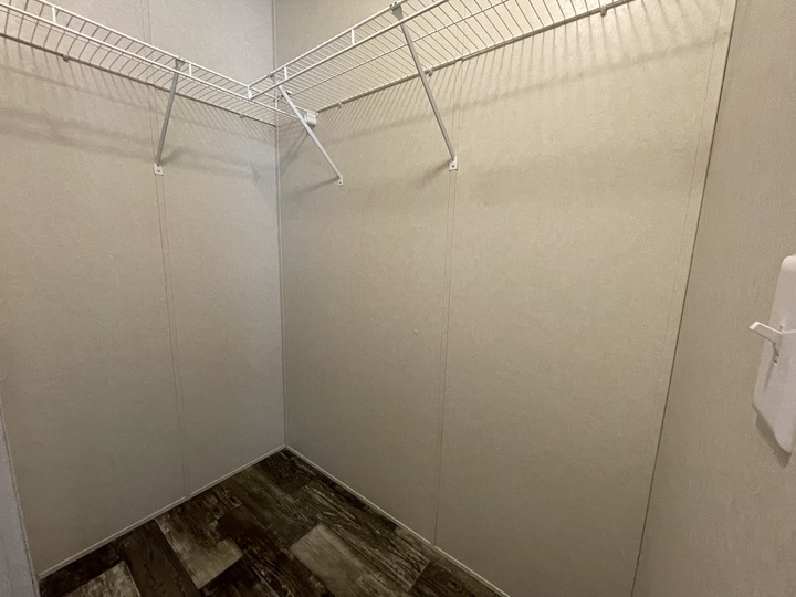 Empty walk-in closet with beige walls, dark wood-patterned floor, and white wire shelves along two walls. There is a small white fixture on the right wall. The space is well-lit.