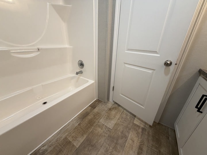 A clean bathroom features a white bathtub with a built-in shelf and faucet, a closed white door, light wood-look flooring, and part of a white vanity with black handles.
