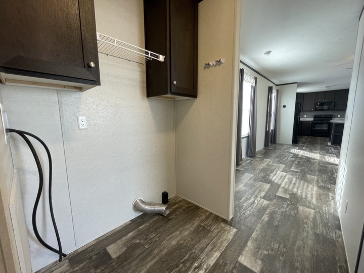 Empty laundry area with washer and dryer hookups, wire shelf, and dark cabinets; view extends into a kitchen with dark cabinetry and appliances, and natural light from windows along the right wall.
