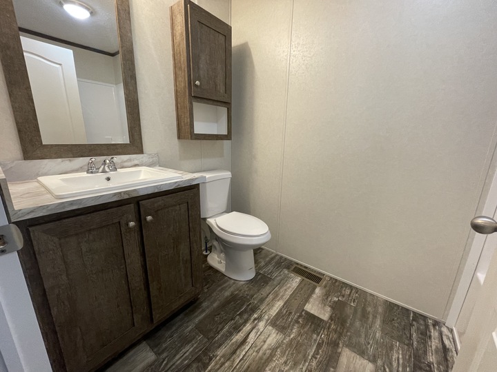 A bathroom with a wooden vanity and sink, a wall-mounted cabinet with mirror, a white toilet, and gray wood-patterned flooring. The walls are light-colored and plain.