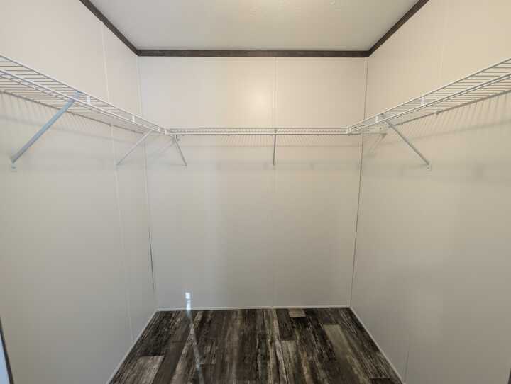 Empty walk-in closet with light-colored walls, dark trim, and two long white wire shelves along both sides. The floor has a wood-like pattern in shades of gray and brown.
