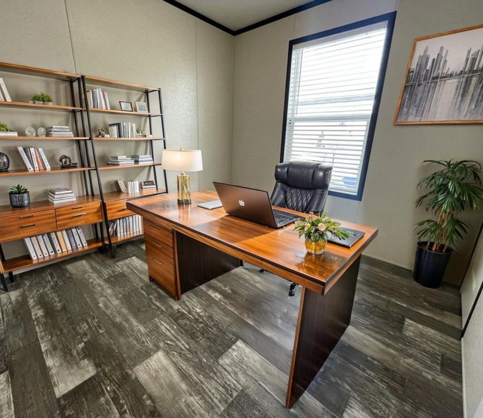 A modern office with a wooden desk, laptop, lamp, and chair. There are bookshelves with books and decor, a potted plant, large window with blinds, and a cityscape artwork on the wall. The floor has a dark wood pattern.