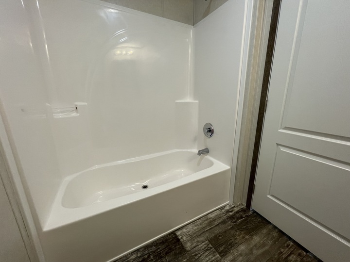 A clean, white bathtub with a built-in shower in a bathroom with white walls, a closed white door, and wood-patterned flooring. The tub has a chrome faucet and a small shelf for toiletries.