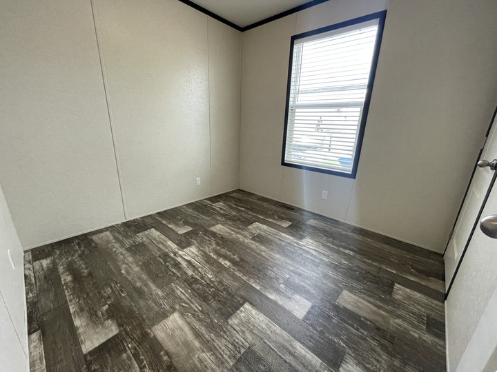 An empty room with light-colored walls, a large window with blinds, and dark wood-patterned flooring. Natural light comes through the window, and the room appears clean and unfurnished.
