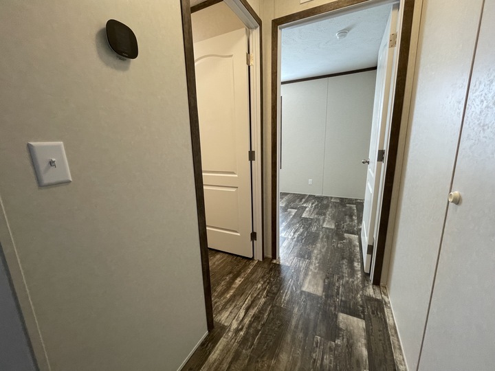 A hallway with wood-patterned flooring, cream-colored walls, and several open white doors leading to adjacent rooms. A light switch and a small black device are mounted on the left wall.