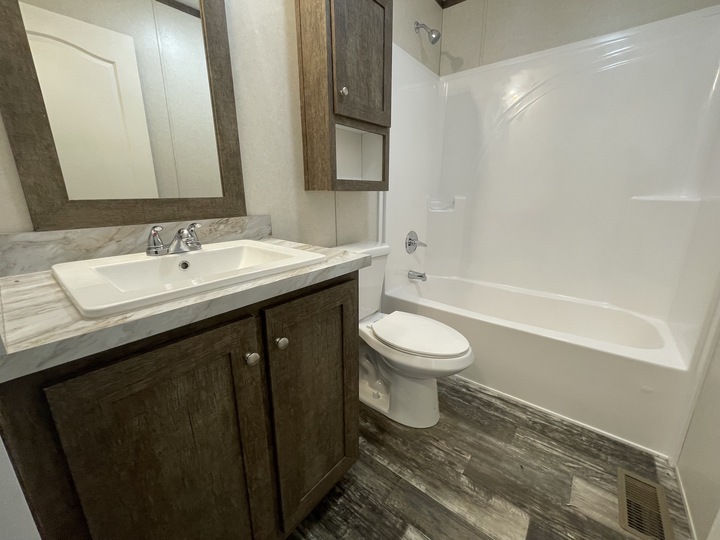 A bathroom with a wood-framed mirror, marble countertop sink, cabinet, toilet, and white shower-tub combo. The floor has a wood-like finish and there is a wall vent near the toilet.