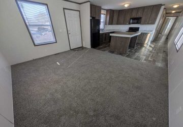 A spacious room with gray carpet, connected to a modern kitchen featuring dark wood cabinets, an island, black appliances, and a hallway leading to other areas; sunlight streams through several windows.