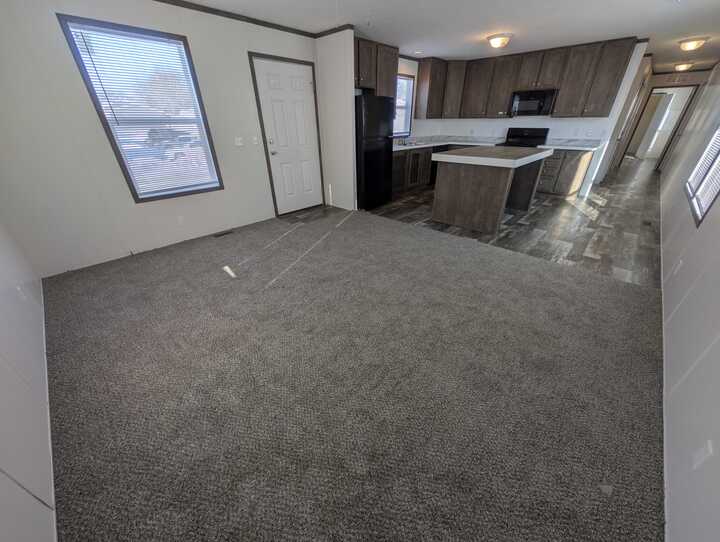 A spacious room with gray carpet, connected to a modern kitchen featuring dark wood cabinets, an island, black appliances, and a hallway leading to other areas; sunlight streams through several windows.