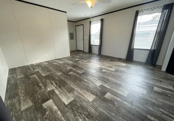 A spacious, empty room with dark wood-patterned flooring, light-colored walls, two large windows with dark curtains, a ceiling fan with a light, and a closed white door. Natural light brightens the space.