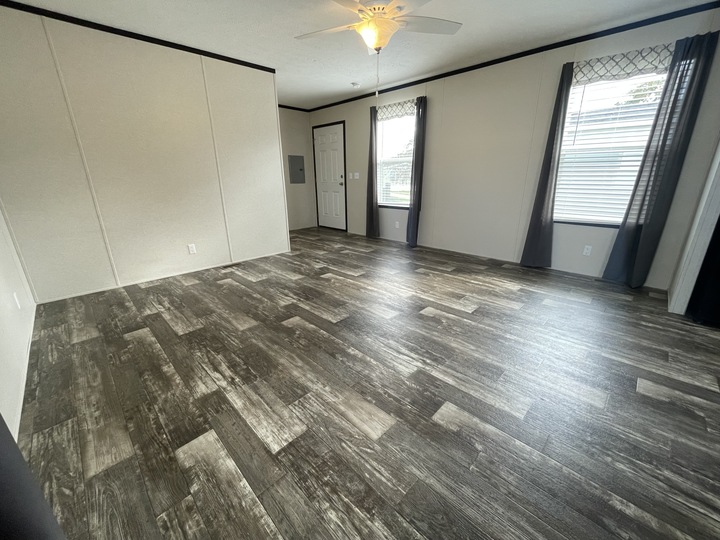 A spacious, empty room with dark wood-patterned flooring, light-colored walls, two large windows with dark curtains, a ceiling fan with a light, and a closed white door. Natural light brightens the space.