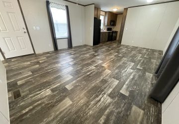 A spacious, empty room with wood-patterned vinyl flooring, beige walls, large windows with dark curtains, and an open view into a small kitchen with dark cabinets and black appliances.