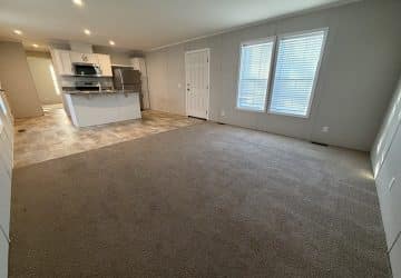 Open living area with beige carpet, large windows with blinds, and an adjoining kitchen featuring white cabinets, stainless steel appliances, and tile flooring. The room is brightly lit with recessed ceiling lights.