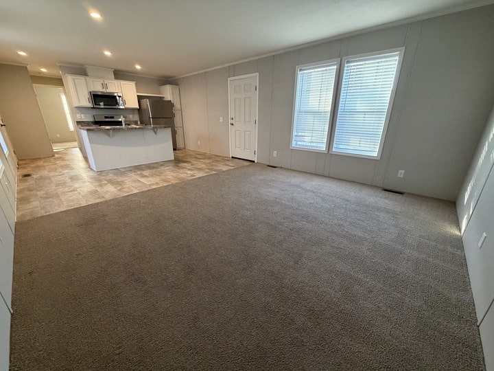 Open living area with beige carpet, large windows with blinds, and an adjoining kitchen featuring white cabinets, stainless steel appliances, and tile flooring. The room is brightly lit with recessed ceiling lights.