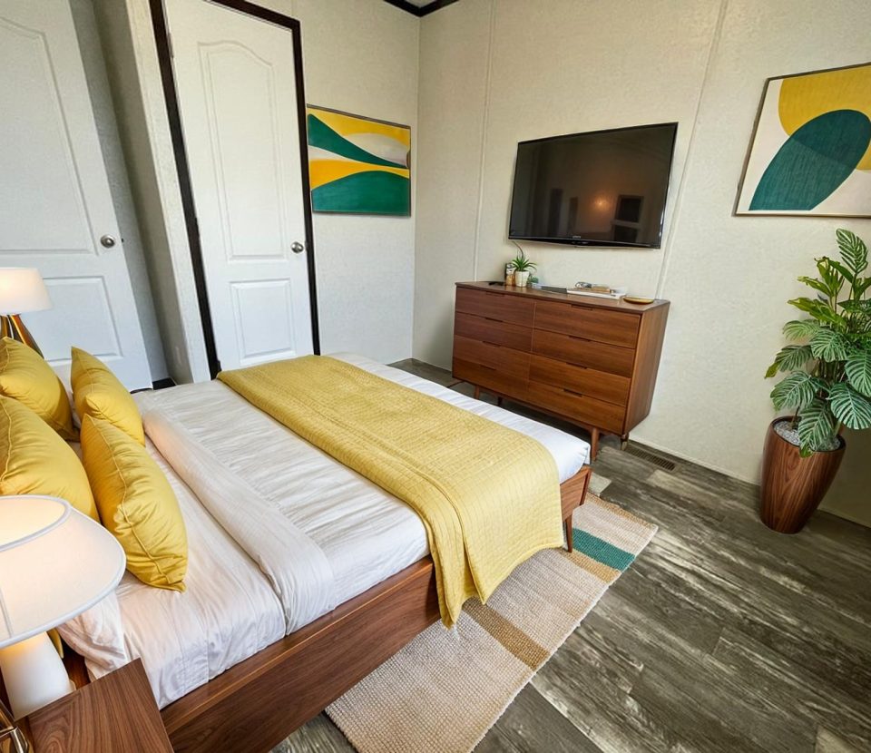 Modern bedroom with a wooden bed, yellow and white bedding, a wooden dresser, a wall-mounted TV, abstract art on the walls, a potted plant, and two white doors. The room has a cozy, tidy feel.