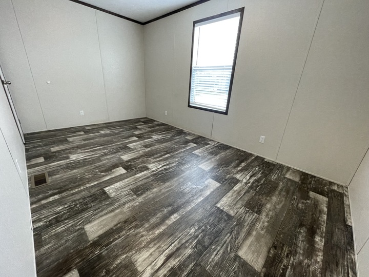 Empty room with light beige walls, dark wood-patterned vinyl flooring, a single window with blinds, white electrical outlets, and a closed door partially visible on the left.