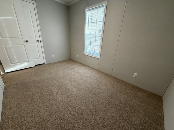 An empty room with beige carpet, light gray walls, a white door with chrome knobs, and a window with white blinds letting in natural light.