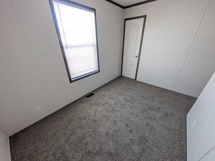 A small, empty room with light-colored walls, a window with blinds, gray carpet flooring, and a closed white door.