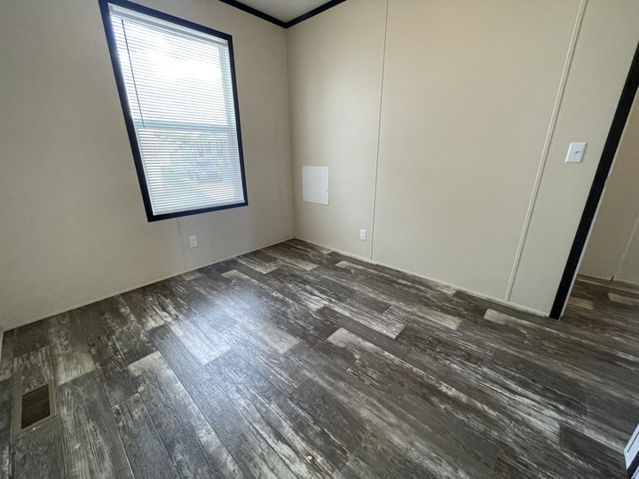 An empty room with beige walls, a large window with blinds, wood-patterned vinyl flooring, and a white wall vent. Light from the window brightens the simple, unfurnished space.