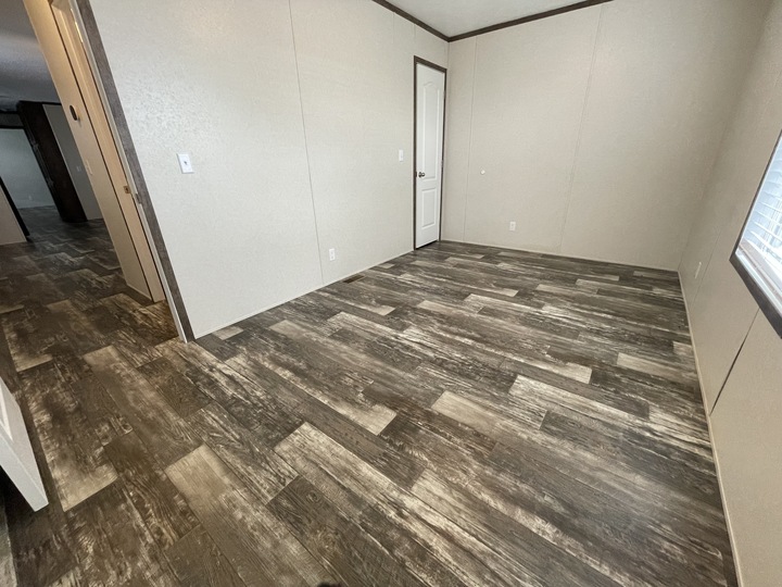 Empty room with light beige walls, dark brown trim, wood-look laminate flooring, a white door, and a window letting in natural light. The room connects to a hallway with matching flooring.