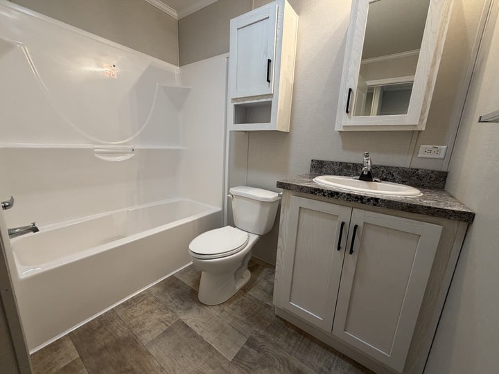 A clean, modern bathroom with a white bathtub, toilet, and sink. The sink sits in a speckled countertop with white cabinets. There is a wall-mounted cabinet above the toilet and neutral-colored walls and floor.